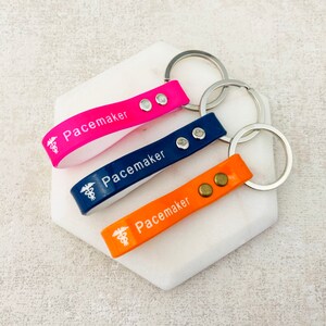 Könnte beinhalten: Drei Schlüsselanhänger mit einem medizinischen Symbol und dem Wort "Pacemaker" darauf gedruckt. Die Schlüsselanhänger sind rosa, blau und orange.