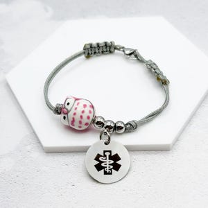 May include: Gray braided bracelet with a pink and white owl charm and a silver medical symbol charm.