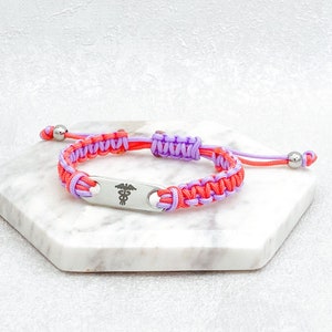 May include: A pink and orange braided bracelet with a silver medical symbol charm. The bracelet is adjustable with a sliding knot.