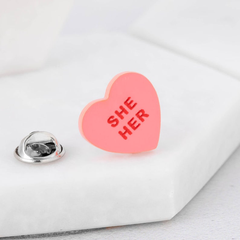 Close-up of She/Her pronoun pin with hand-painted lettering on acrylic heart design