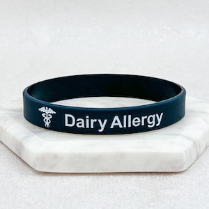 Peut inclure: Bracelet en silicone noir avec le texte blanc "Allergie aux produits laitiers" et un symbole médical blanc.