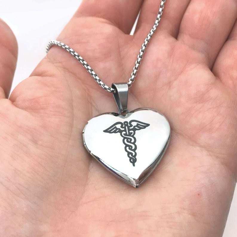 May include: A silver heart-shaped locket pendant with a medical caduceus symbol. The pendant hangs from a delicate chain. The locket is held in a hand.