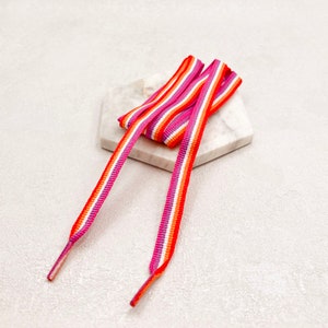 May include: A pair of shoelaces with pink, orange and white stripes. The shoelaces are laid out on a white marble surface.