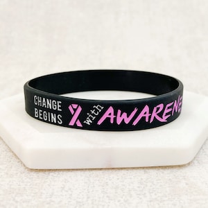 May include: Black silicone bracelet with the text "Change Begins with Awareness" in pink lettering and a pink ribbon symbol.