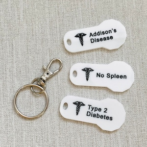 May include: Three white keychains with a medical symbol and text. The keychains read "Addison's Disease", "No Spleen", and "Type 2 Diabetes".