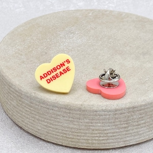 May include: A yellow heart-shaped pin with the text "ADDISON'S DISEASE" in red letters. The back of the pin is pink.