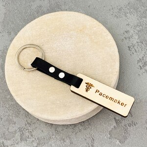 Könnte beinhalten: Holzschlüsselanhänger mit schwarzem Lederband und silbernem Schlüsselring. Der Schlüsselanhänger hat ein medizinisches Symbol und den Schriftzug "Pacemaker" eingraviert.