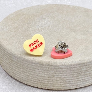 May include: A heart-shaped pin with the text "PACE MAKER" in red on a yellow background. The pin has a silver backing and a pink heart-shaped base. The pin is on a light gray surface.