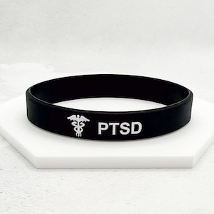 May include: Black rubber wristband with the word "PTSD" in white lettering. The wristband also features a white medical symbol. The wristband is displayed on a white hexagonal surface.