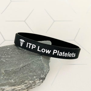 May include: Black silicone wristband with white text and a medical symbol. The text reads "ITP Low Platelets". The wristband is resting on a gray rock against a white background with a geometric pattern. A medical alert bracelet.