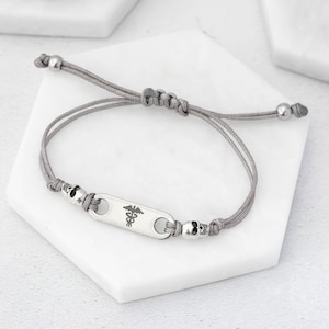 May include: Gray adjustable bracelet with a silver rectangular charm featuring the medical symbol, the Rod of Asclepius. The charm is accented with two silver skull beads.