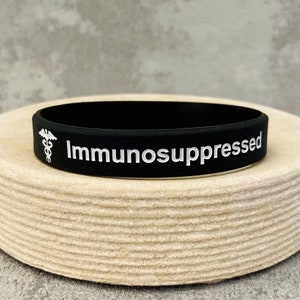 Puede incluir: Pulsera de silicona negra con el texto "Inmunodeprimido" y un s&iacute;mbolo m&eacute;dico blanco.