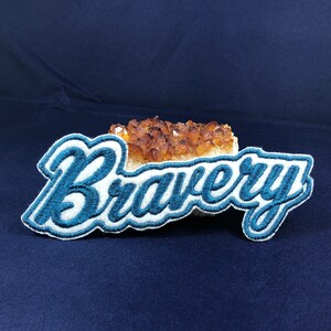 May include: A blue and white embroidered patch with the word "Bravery" in a cursive font. The patch is sitting on a piece of brown crystal.