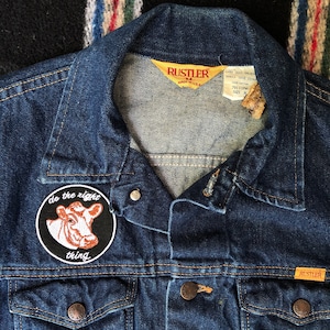 May include: A dark blue denim jacket with a cow patch that says "do the right thing" on the left chest. The jacket has a Rustler brand label on the right chest and a Rustler brand tag on the inside of the collar.