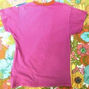 May include: A pink t-shirt with an orange ribbed collar. The shirt is lying flat on a floral fabric background.