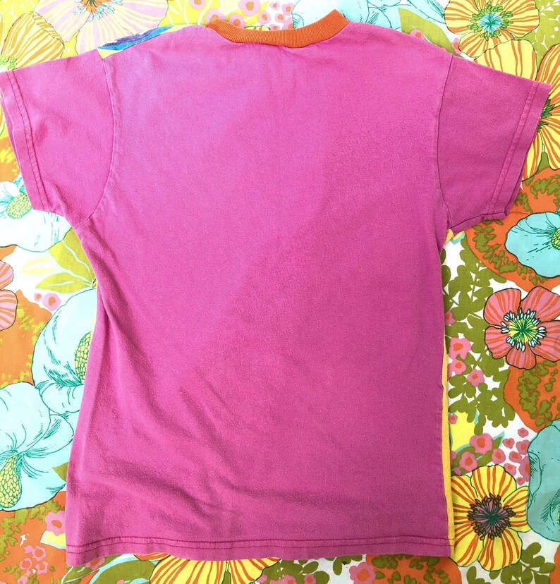 May include: A pink t-shirt with an orange ribbed collar. The shirt is lying flat on a floral fabric background.