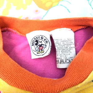 May include: A pink, orange and yellow striped t-shirt with a Mickey Mouse & Co. tag. The tag features a picture of Mickey Mouse and the text "Assembled in Panama, US Components". The tag also includes the size "SC7/B".