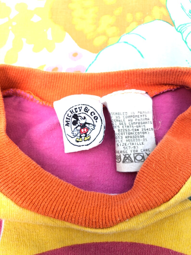May include: A pink, orange and yellow striped t-shirt with a Mickey Mouse & Co. tag. The tag features a picture of Mickey Mouse and the text "Assembled in Panama, US Components". The tag also includes the size "SC7/B".