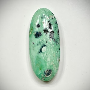 May include: Oval-shaped gemstone with a light green hue and dark, speckled inclusions. The polished stone has a smooth surface and a natural, organic appearance. The gemstone is set against a white background.