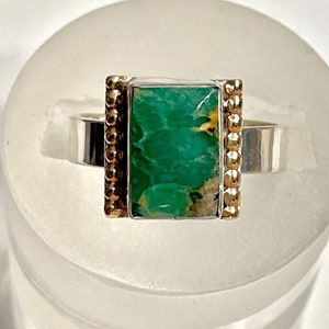 May include: A silver ring with a green turquoise stone set in a gold-toned bezel. The ring has a beaded edge.