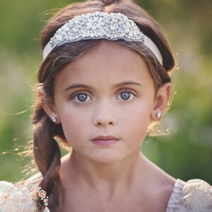 May include: A young girl with blue eyes and a braided hairstyle is wearing a white headband adorned with sparkling crystals. She also has small, round, silver earrings. The background is a soft, blurred green.