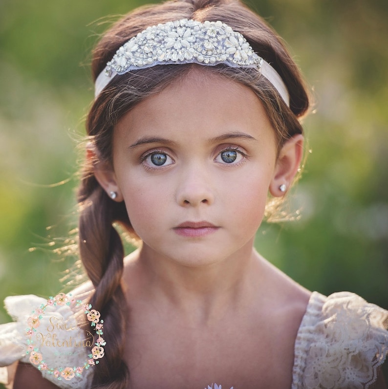 May include: A young girl with blue eyes and a braided hairstyle is wearing a white headband adorned with sparkling crystals. The headband sits atop her head, and she is wearing a cream-colored dress. The background is a soft, blurred green.