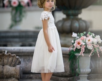 May include: A young girl wearing a white tulle dress with lace detailing and a flower crown. She is standing in front of a fountain and a potted plant.
