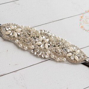 May include: An elegant headband featuring a central design of sparkling rhinestones, faux pearls, and clear crystals. The embellishment is attached to a black elastic band. The headband is suitable for special occasions.