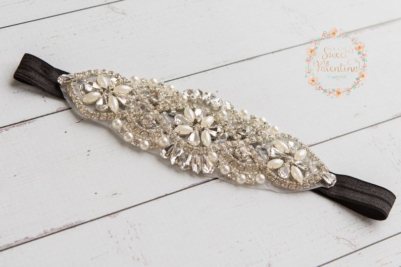 May include: An elegant headband featuring a central design of sparkling rhinestones, faux pearls, and clear crystals. The embellishment is attached to a black elastic band. The headband is suitable for special occasions.
