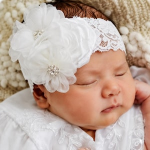 May include: A white lace headband featuring two large, layered fabric flowers. Each flower is accented with a central cluster of small white pearls and a clear, faceted rhinestone. The headband is made of a delicate, floral-patterned white lace.