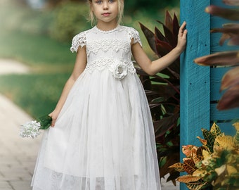 May include: A young girl wearing a white tulle dress with lace detailing and a flower accent at the waist. She is holding a bouquet of white flowers and is standing next to a blue wooden fence.