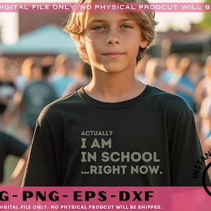 May include: A boy wearing a black t-shirt with the text "Actually I am in school...right now." printed on it. The boy is standing in front of a blurred background of people.