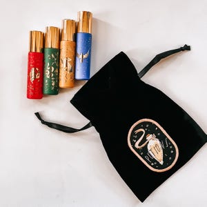 May include: Four cylindrical roller bottles in red, green, yellow, and blue, each featuring gold metallic caps and intricate gold foil designs. Beside them is a black velvet drawstring pouch with an embroidered patch depicting a potion bottle.