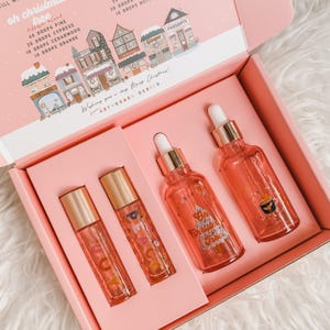 May include: A pink gift box containing two rollerball bottles labeled JOY and PEACE, and two dropper bottles labeled Oh Christmas Tree and a coffee cup graphic. The box lid features a winter village illustration and essential oil blend recipes.