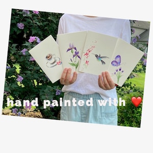 K&ouml;nnte beinhalten: Vier handgemalte Karten mit einem Kolibri, einem Schmetterling, einer Irisbl&uuml;te und einer Tasse Kaffee. Die Karten werden von einer Person vor einem gr&uuml;nen Busch gehalten.