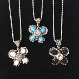 May include: Three silver butterfly necklaces with different colored stones. The top necklace has turquoise stones, the middle necklace has white stones, and the bottom necklace has black stones. Each butterfly has a bullet casing in the center with the text "9mm Luger" engraved on it.