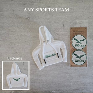 Puede incluir: Sudadera blanca con capucha con un logotipo verde "Eagles" en el bolsillo delantero. Dos posavasos redondos para coche con el mismo logotipo se muestran sobre un fondo de madera. El texto "Car Coasters" está impreso en la madera.