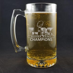 May include: A clear glass beer mug filled with a light amber liquid. The mug has a handle and a decorative base. The front of the mug features the text "SUPER BOWL CHAMPIONS" with a graphic of a trophy, trees, and a bridge.