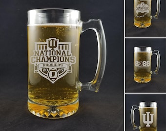May include: A clear glass beer mug filled with beer, featuring a handle and a decorative base. The mug is etched with "NATIONAL CHAMPIONS HOOSIERS 20 25". Additional mugs with different designs are also shown.