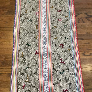 May include: A white woven tapestry with a geometric pattern in black, red, green, and yellow. The tapestry has a colorful border with stripes of red, blue, yellow, and green.