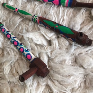 May include: Two wooden pipes with colorful floral designs painted on them. The pipes are made of dark brown wood and have a small bowl at the end. The pipes are lying on a white fluffy surface.