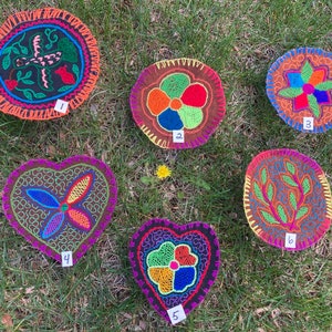 Pode incluir: Cinco círculos e corações de tecido bordados com desenhos florais coloridos. Os círculos são de cor marrom, laranja e preto com bordados verdes, vermelhos, azuis e amarelos. Os corações são de cor marrom com bordados verdes, vermelhos, azuis e amarelos. Cada peça é numerada de 1 a 5.