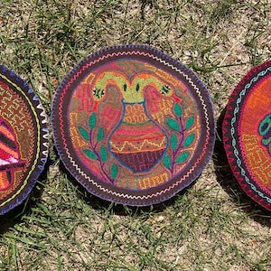 May include: Three colorful, round embroidered patches with nature-themed designs. One features a vibrant owl, another a vase with birds, and the last a frog. Each patch has a dark border and is set against a grassy background.