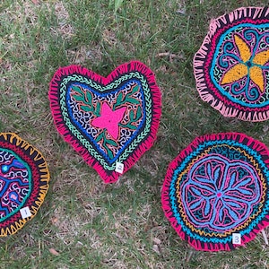 Op de afbeelding: Vier geborduurde stoffen cirkels en een hartvormig stoffen stuk. De cirkels zijn in verschillende kleuren, waaronder roze, geel, blauw en groen. Het hart is roze, blauw en groen. Elk stuk heeft een ander ontwerp en is genummerd van 1 tot 4.