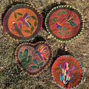 Pode incluir: Quatro bases para copos bordadas com desenhos coloridos. Duas são redondas, uma em forma de coração e outra com uma rã. As bases para copos têm centros castanhos com imagens costuradas de beija-flores, cactos e uma rã, com costuras coloridas e bordas decorativas.