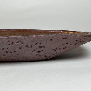 May include: A shallow, elongated ceramic dish with a dark, porous, and textured exterior surface in shades of brown. The interior of the vessel features a smooth, lighter brown finish with subtle speckling, showcasing an irregular, organic rim.