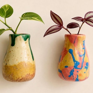 May include: Two ceramic wall vases. The left vase has a cream, yellow, and green drip glaze with a green plant. The right vase has a pink, yellow, and blue splatter paint finish with a purple and silver striped plant. Both have a rounded shape.