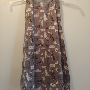 May include: A gray Star Wars themed apron with a repeating pattern of Rey, Kylo Ren, and the Star Wars logo. The apron has a halter neck style and is made of a soft, comfortable fabric.