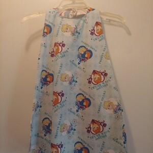 May include: A light blue sleeveless dress with a white satin neckline. The dress is covered in a repeating pattern of Frozen characters, snowflakes, and the text "Frozen Heart", "Strong Bond", and "Never Give Up".