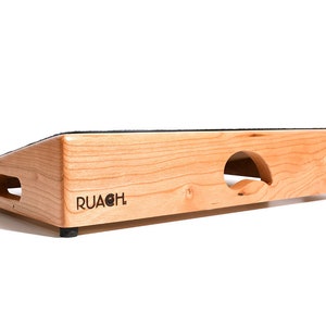 Może przedstawiać: Drewniany pedalboard z czarną tkaniną na górze i wycięciem na kable. Pedalboard jest pochylony i ma wygrawerowane słowo "RUACH" z przodu.